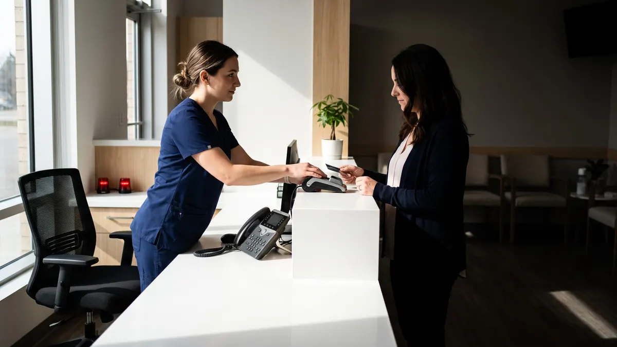 A dental receptionist helping a patient pay at the front desk, the office phone beside her and the second chair empty.