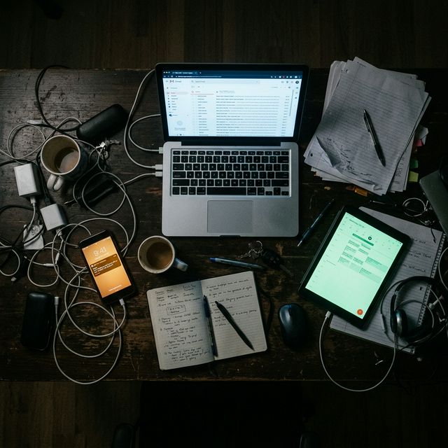 A dark desk with laptop, phone, tablet, and notebooks spread apart , every device a different communication island with no connection between them.